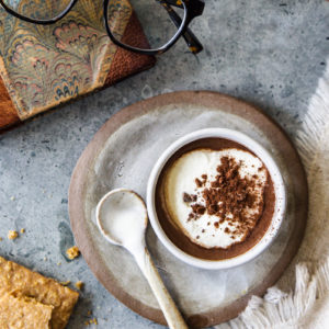 a rustic pottery cup of drinking chocolate on a saucer with a ceramic spoon, with whipped cream and shaved chocolate on a stone tabletop with cookies, a marbled book and a pair of glasses for a cozy scene