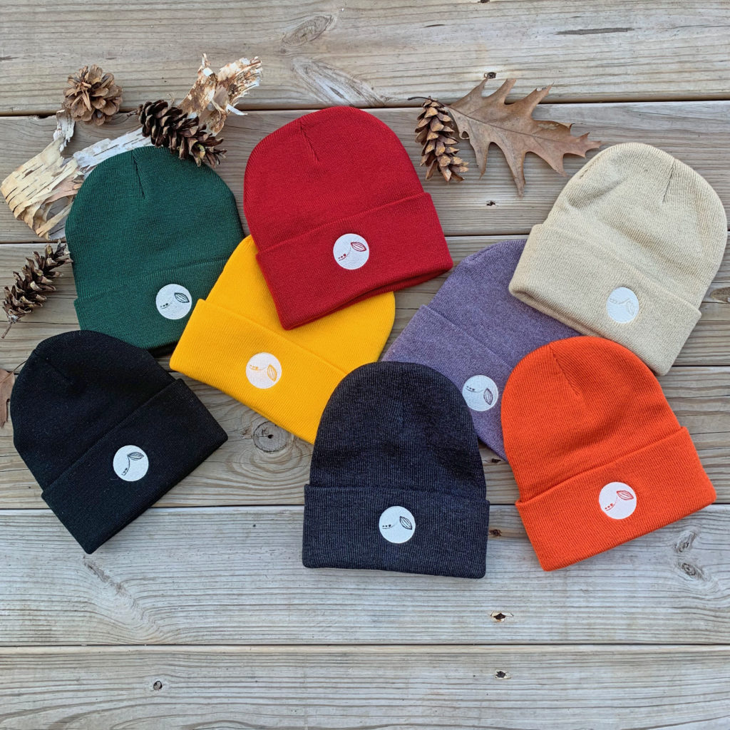an array of knit beanies in a rainbow of earthy colors on a barnyard background with pinecones and birch bark