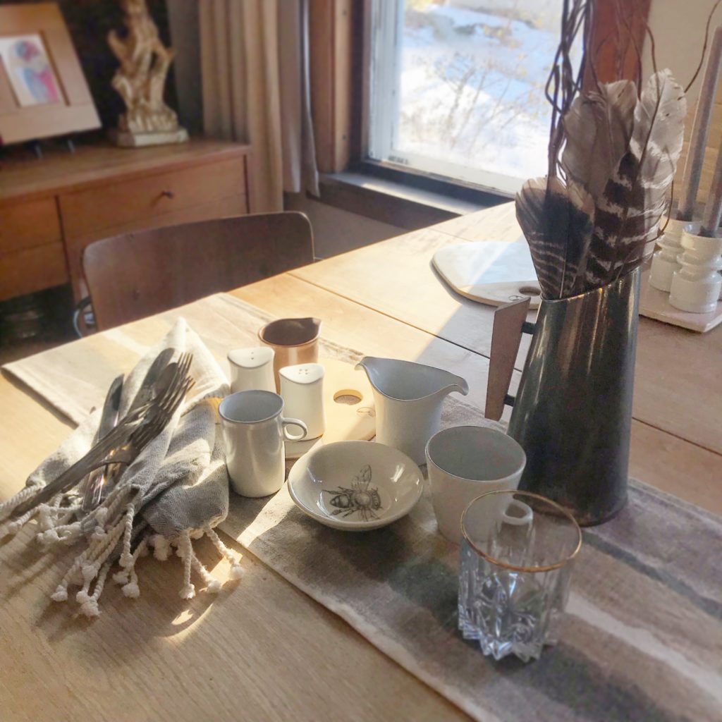 Small white mugs on a wooden table. On the table is also a silver stein with turkey feathers in it