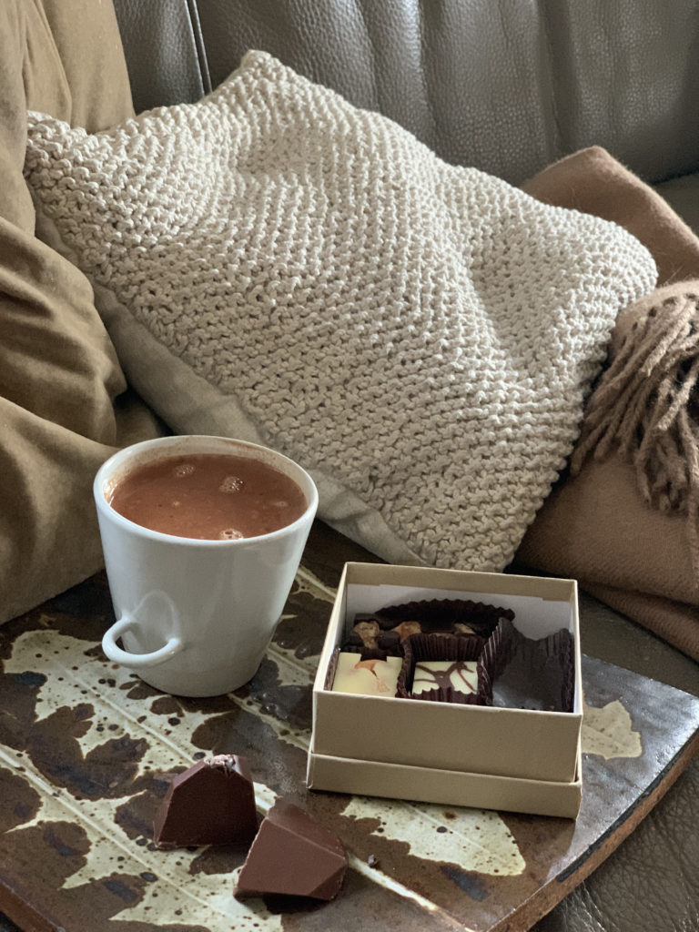 Box of Tavernier chocolates and a mug of sipping chocolate. Behind these is a knit pillow and a blanket