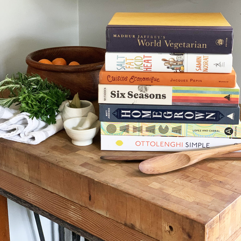 Cutting board topped with mortar and pestle and a collection of cookbooks. Some of the books featured are "Salt, Acid, Fat, Heat," "Six Seasons," "and "Ottolenghi Simple"
