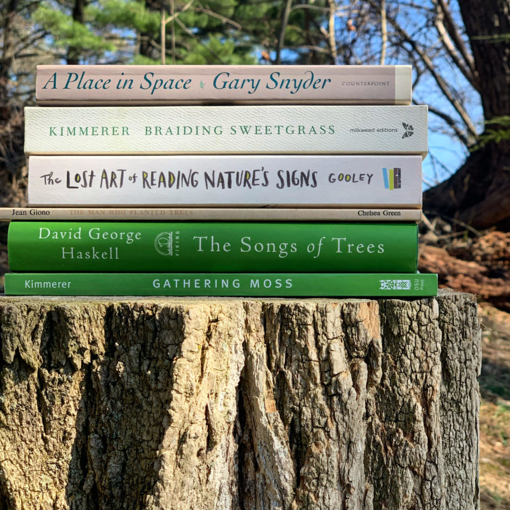 A collection of eco-books stacked atop a tree stump. Some of the books featured are "Place in Space," "The Songs of Trees," and "Gathering Moss"