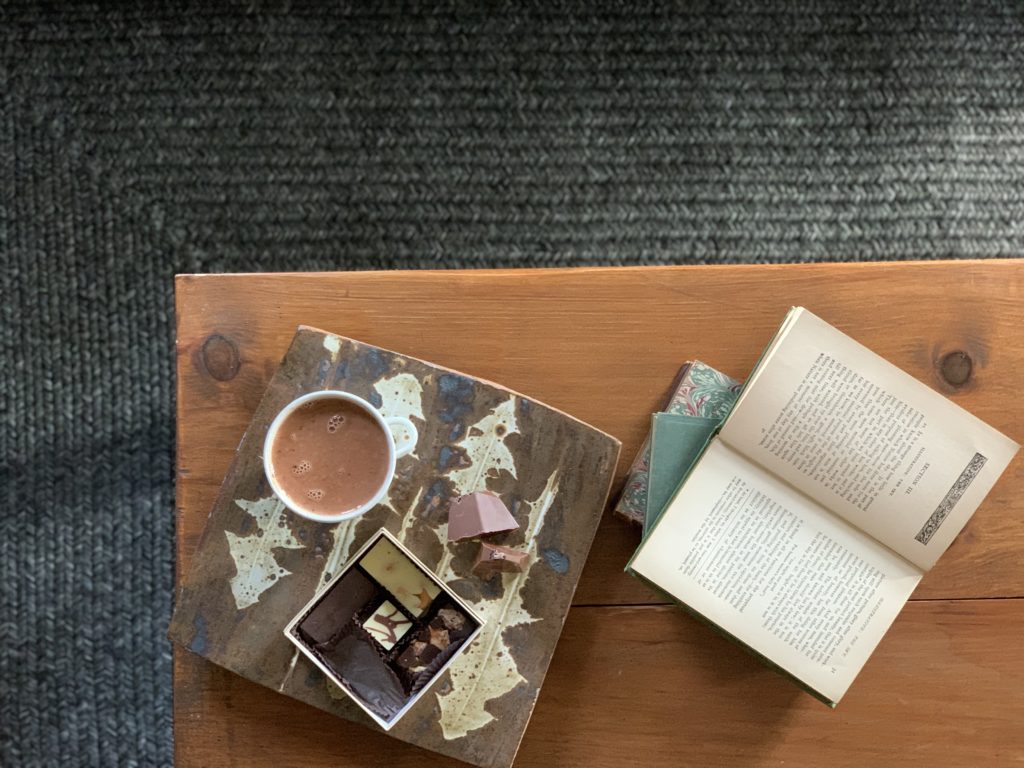 Overhead shot of a box of bonbons and sipping chocolate. Next to these is an open book. All are sitting on a wooden table