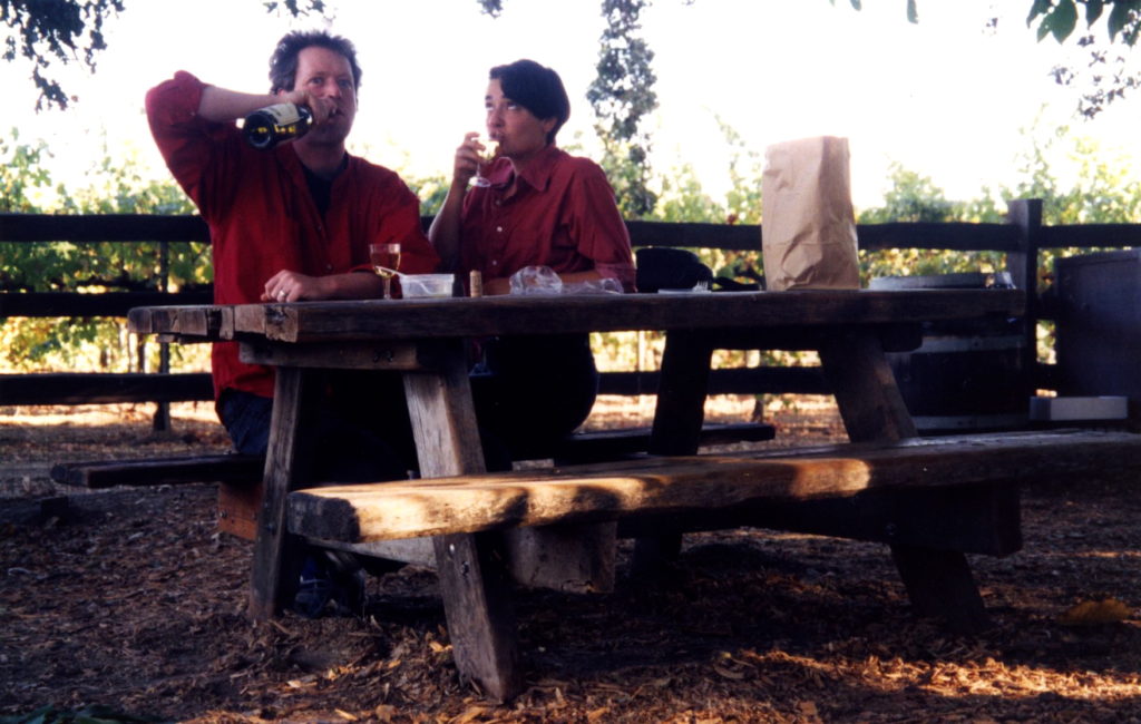 Vintage photo of John and Dar sitting at a picnic table sharing food and a bottle of wine. Dar is sipping the wine and john is sipping from the bottle in a humorous way