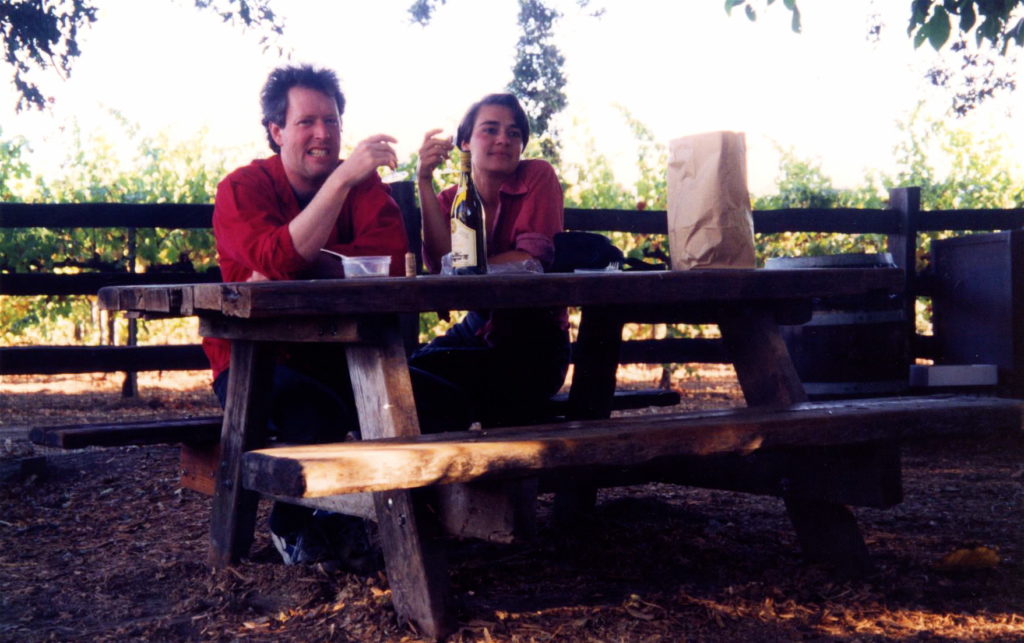 Vintage photo of John and Dar sitting at a picnic table sharing food and a bottle of wine