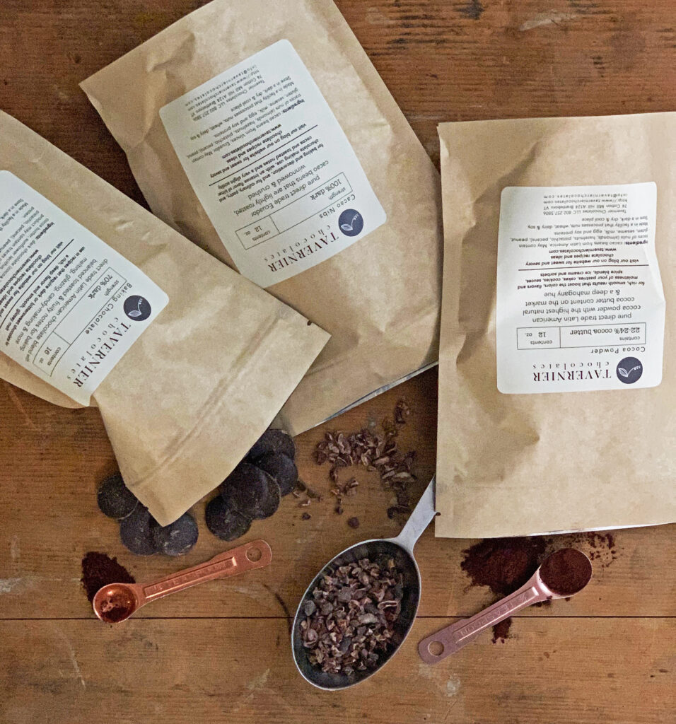 kraft bags of cocoa powder, baking chocolate and cocoa nibs with some spilled onto a wood countertop next to copper measuring spoons and a tin measuring cup