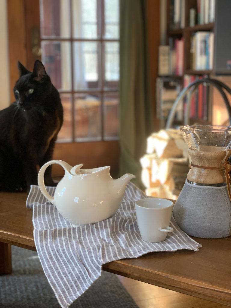 Black cat sitting on a wooden table next to a white teapot and a mug. 