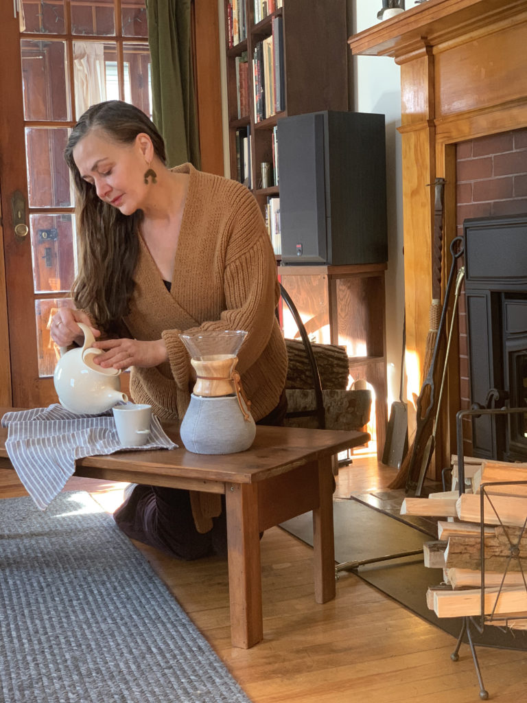 Tar Singer in her home by the fireplace, pouring tea from a teapot.