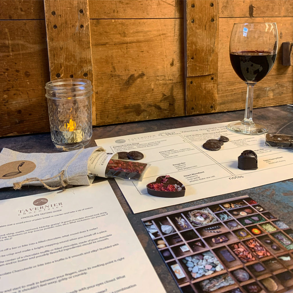 Brochures and menus from Tavernier laid out on a table with a glass of red wine, a mason car with a tealight, and several bonbons.