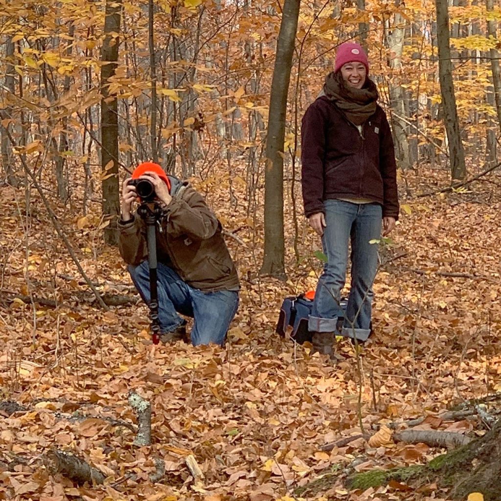 Two filmmakers are standing in the woods during fall. On the left is a man crouching down behind a camera. And on the right is a woman standing and smiling at the camera, wearing a pink beanie.
