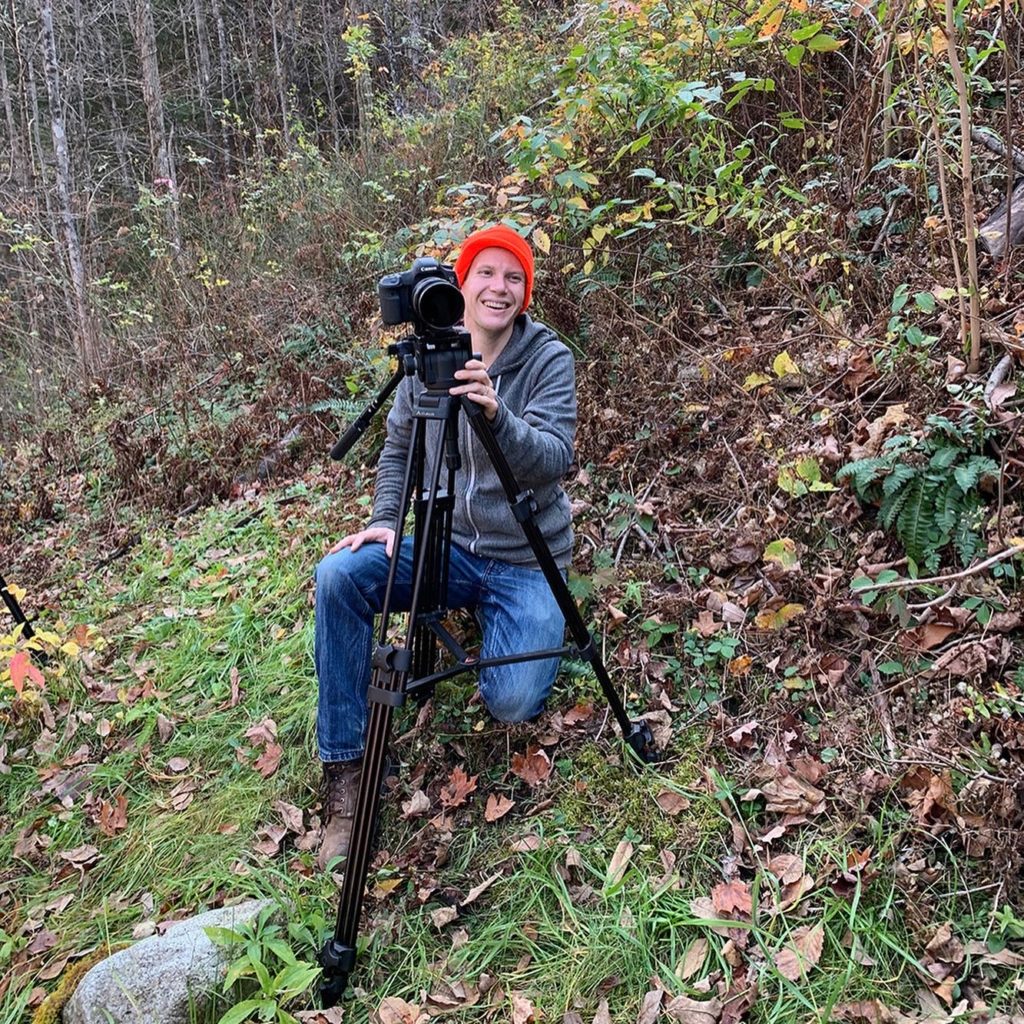 A filmmaker crouches behind his camera which is on a tripod. He's smiling and is on the side of a hill in the woods. He wears a bright orange beanie.