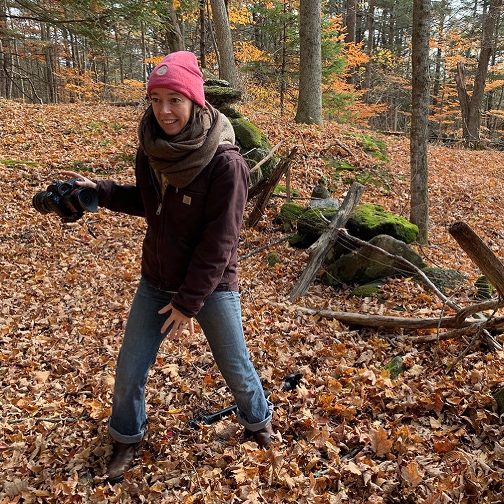 Candid shot of a filmmaker in the woods. The filmmaker is a woman wearing a pink Tavernier beanie, a dark purple carhartt sweatshirt and blue jeans. In her hand, she's holding a large camera.