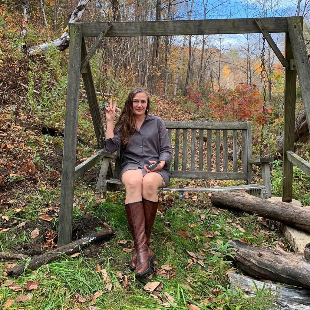 Outside in the fall woods, Dar Singer sits on a wooden rocking chair. She's looking at the camera and holding up a peace sign. She's wearing a grey shirt dress and knee high brown boots.