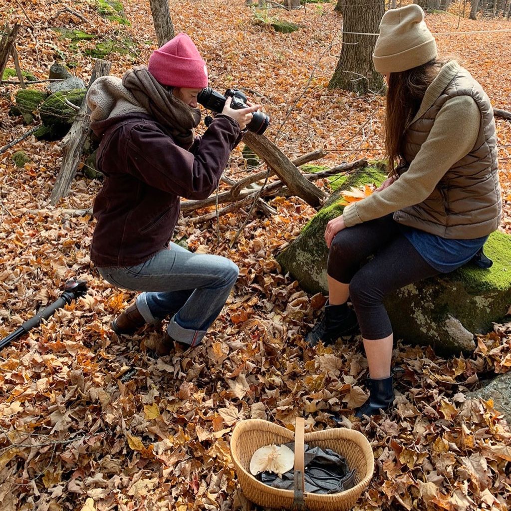 A women wearing a pink beanie and a purple sweatshirt is pointing a camera down at Dar Singer. They are outside in the woods during fall, and Dar is sitting on a moss covered boulder.