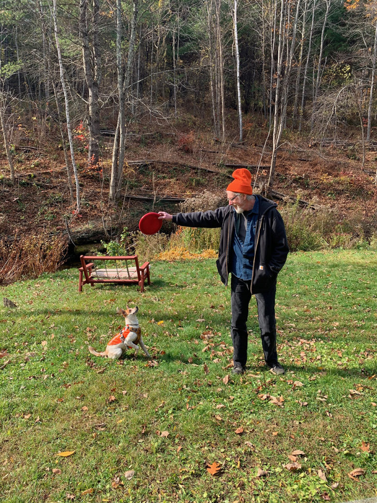 John Singer is outside wearing a jacket and a bright orange beanie. He's holding out a red frisbee for a small white and brown dog wearing an orange vest.