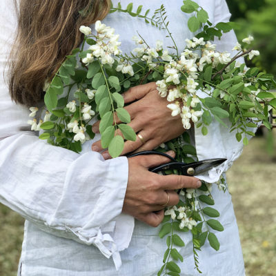 Woman in a white linen shirt is holding a bouquet of small white flowers and green leaves to her chest. In one hand, she holds a pair of shears. We only see her body, not her face.