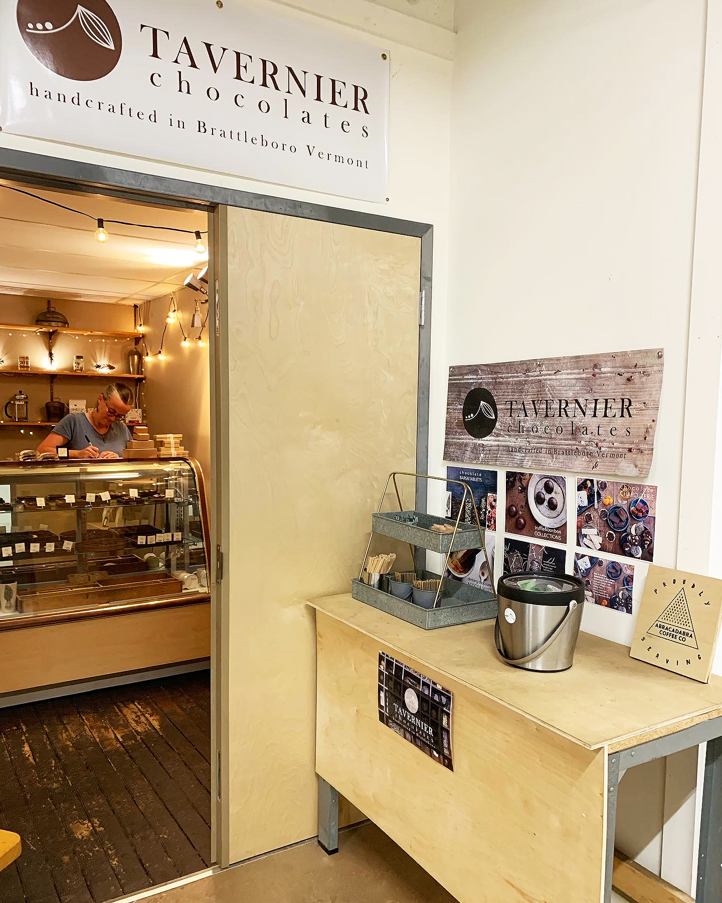 Little shop in an industrial building, door is open and you can see a case with chocolate bonbons, a table outside the door with coffee service, and warm string lights inside.