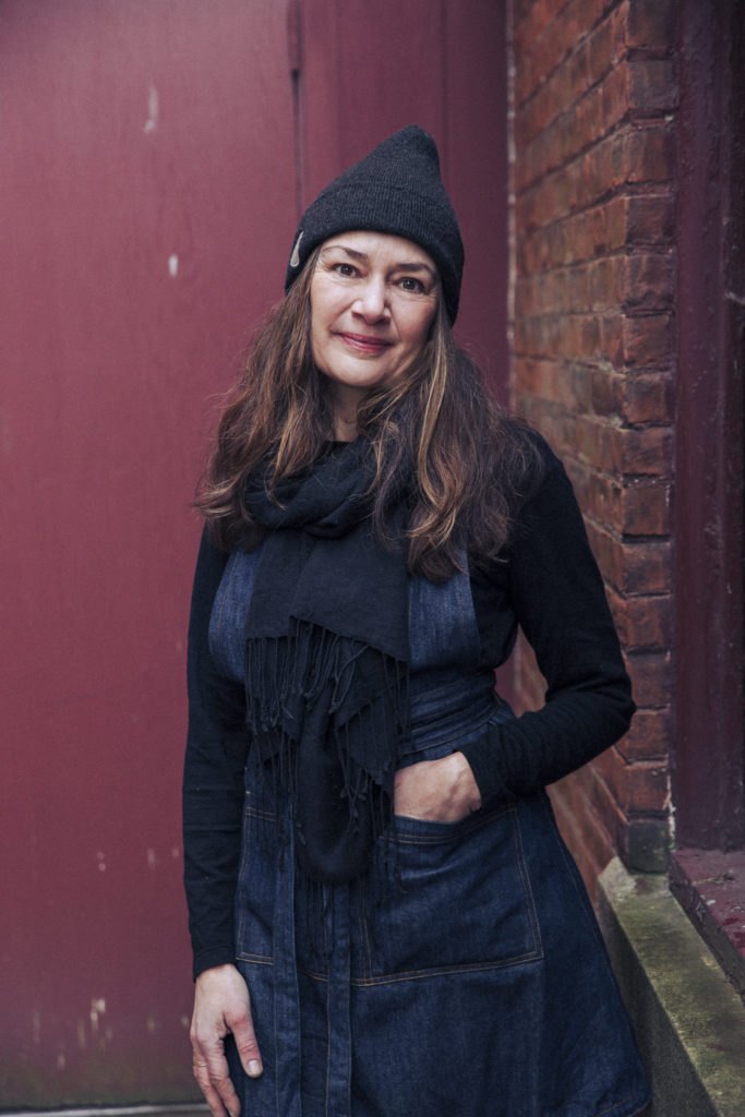 Portrait of chocolatier Dar Tavernier-Singer in a denim apron and dark beanie in front of a red and brick background