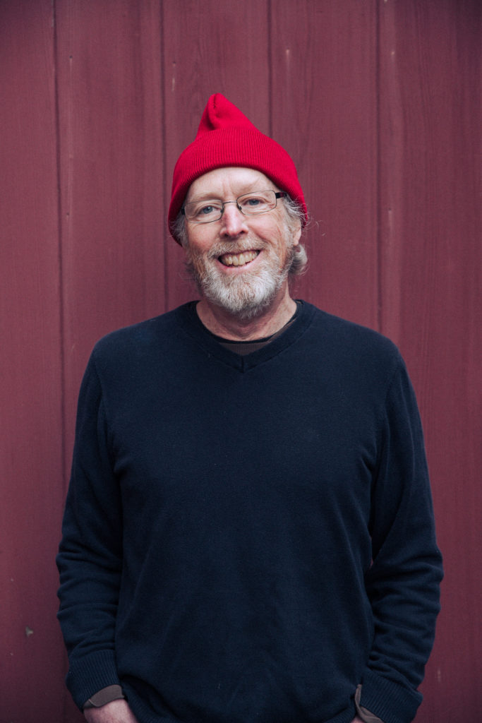 portrait of chocolatier john singer in a dark sweater and red beanie in front of a red background