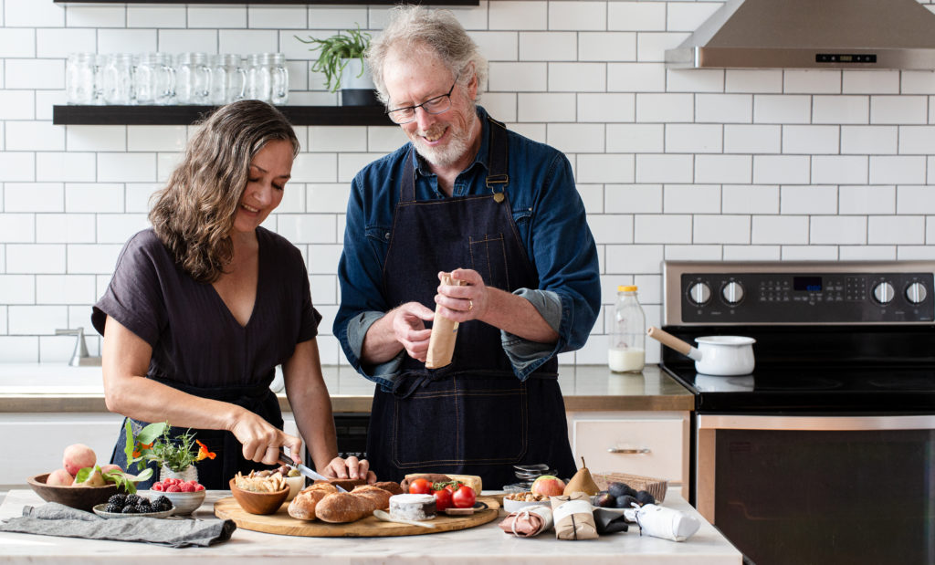 Dar Tavernier and John Singer of Tavernier Chocolates in a white tiled kitchen by a marble kitchen island making a charcuterie board with chocolate, fruit, bread, crackers and herbs