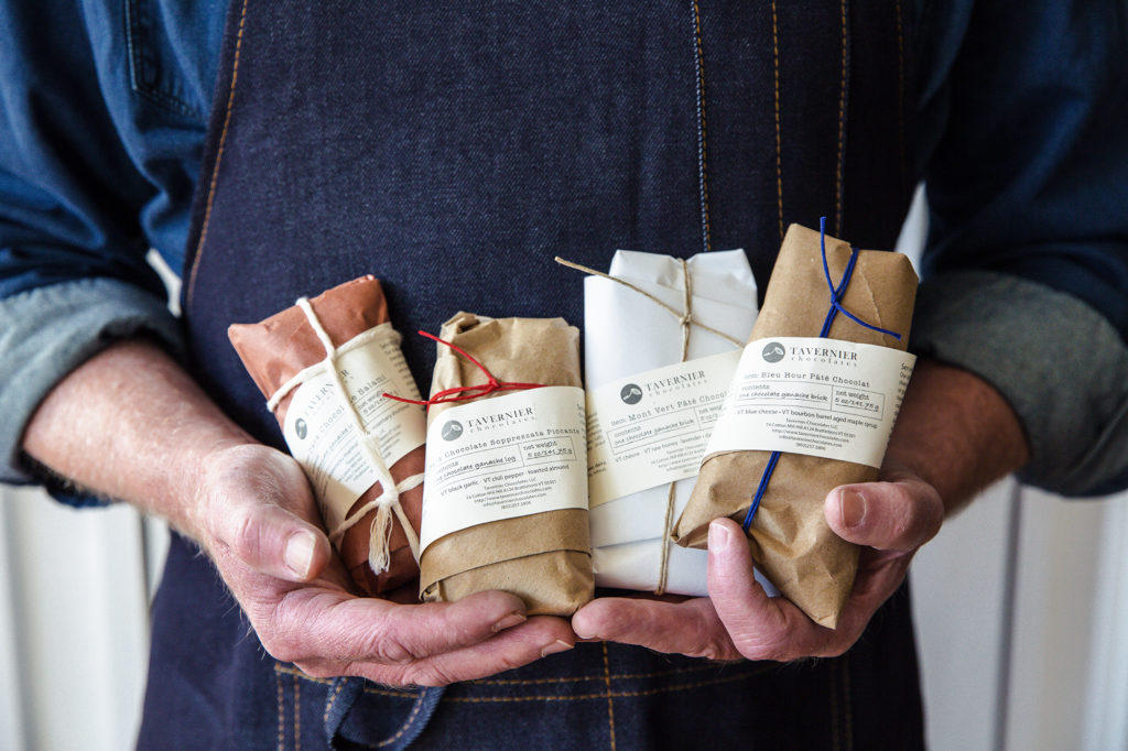 man in a blue chambray shirt and denim apron holding Tavernier Chocolates chocolate charcuterie wrapped in butcher paper and twine to look like real charcuterie