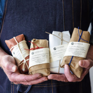 man in a blue chambray shirt and denim apron holding Tavernier Chocolates chocolate charcuterie wrapped in butcher paper and twine to look like real charcuterie