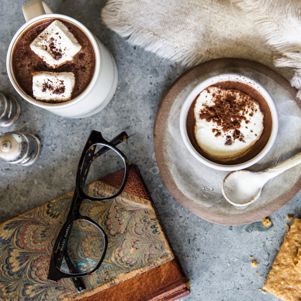 two mugs of hot chocolate one with marshmallows one with whipped cream on a stone table with a marbled cover book, tortoise shell eyeglasses and a linen napkin